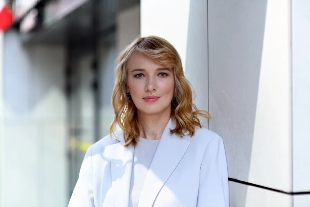 Жінка у білому піджаку стоїть біля стіни