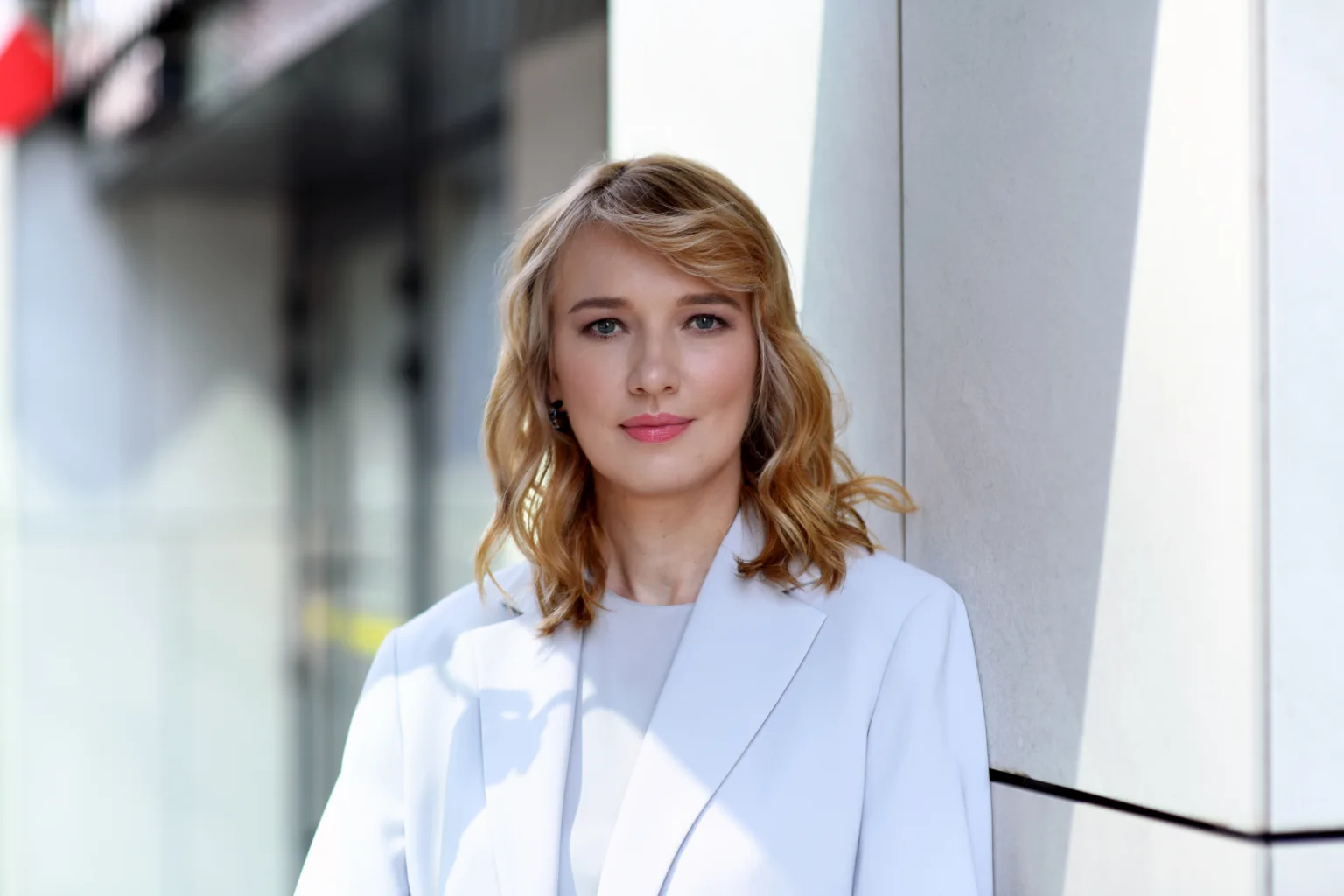 Жінка у білому піджаку стоїть біля стіни