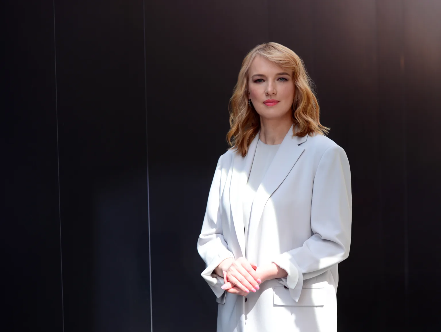 Жінка у світлому костюмі стоїть перед темною стіною