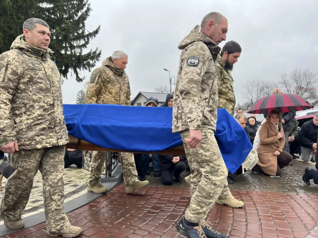 Військові несуть труну, укриту синім прапором