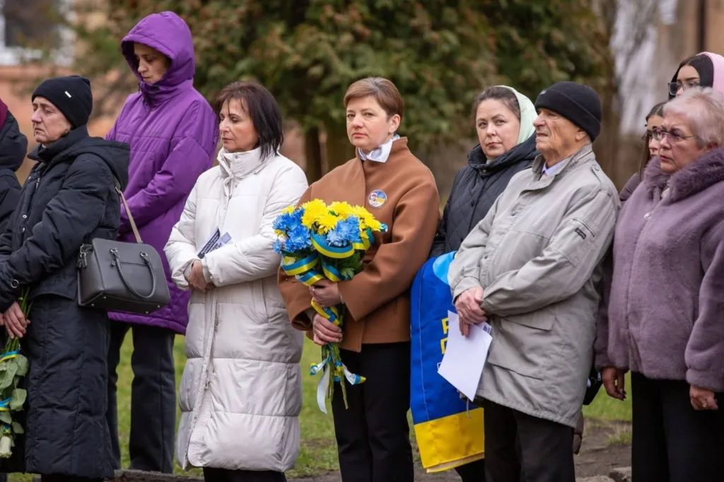 Люди на меморіальному заході з квітами та прапором