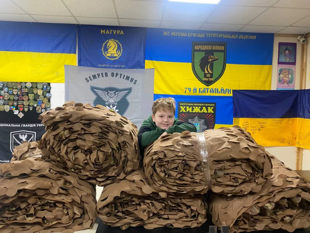 Дитина з маскувальними сітками перед українськими прапорами