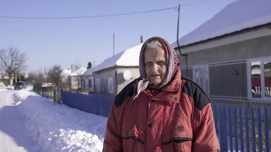 Жінка в червоній куртці стоїть на снігу біля будинку