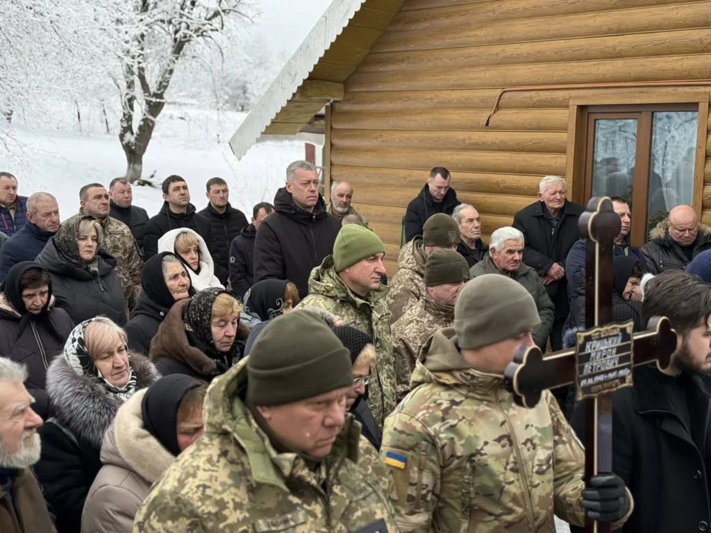 Люди на похороні військового взимку біля дерев'яного дому