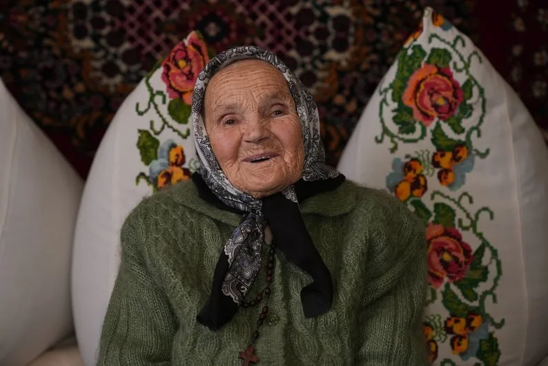 Усміхнена літня жінка в хустці сидить удома