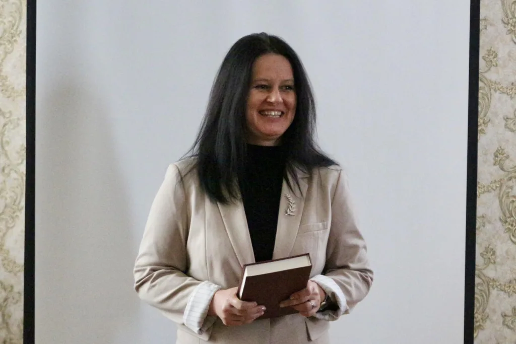 Жінка у піджаку тримає книгу і усміхається