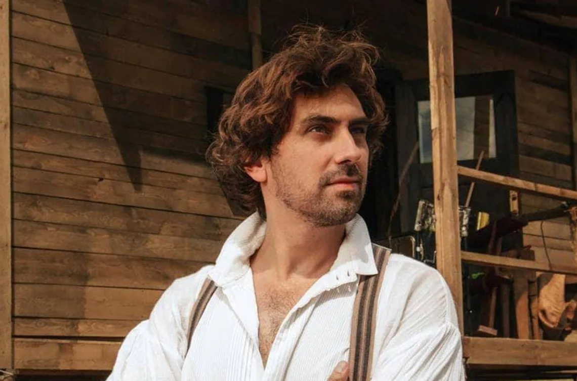 Чоловік у сорочці стоїть біля дерев'яного будинку