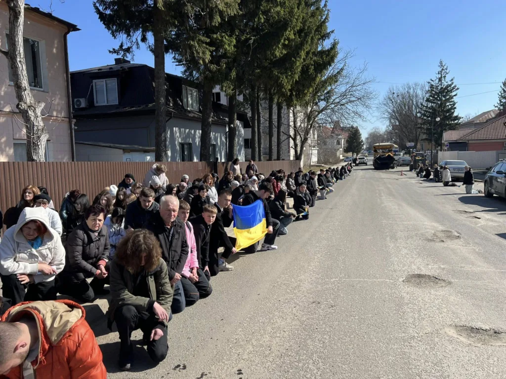 Люди стоять на колінах уздовж дороги з прапором України
