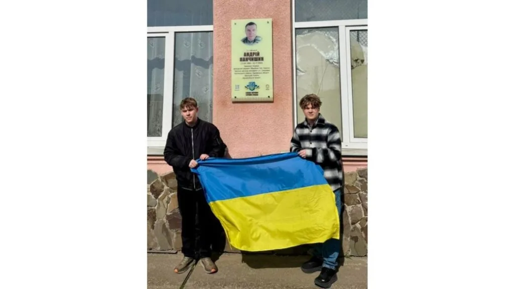 Двоє людей тримають прапор України біля меморіальної дошки.