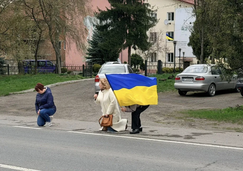 Люди на колінах з українським прапором біля дороги
