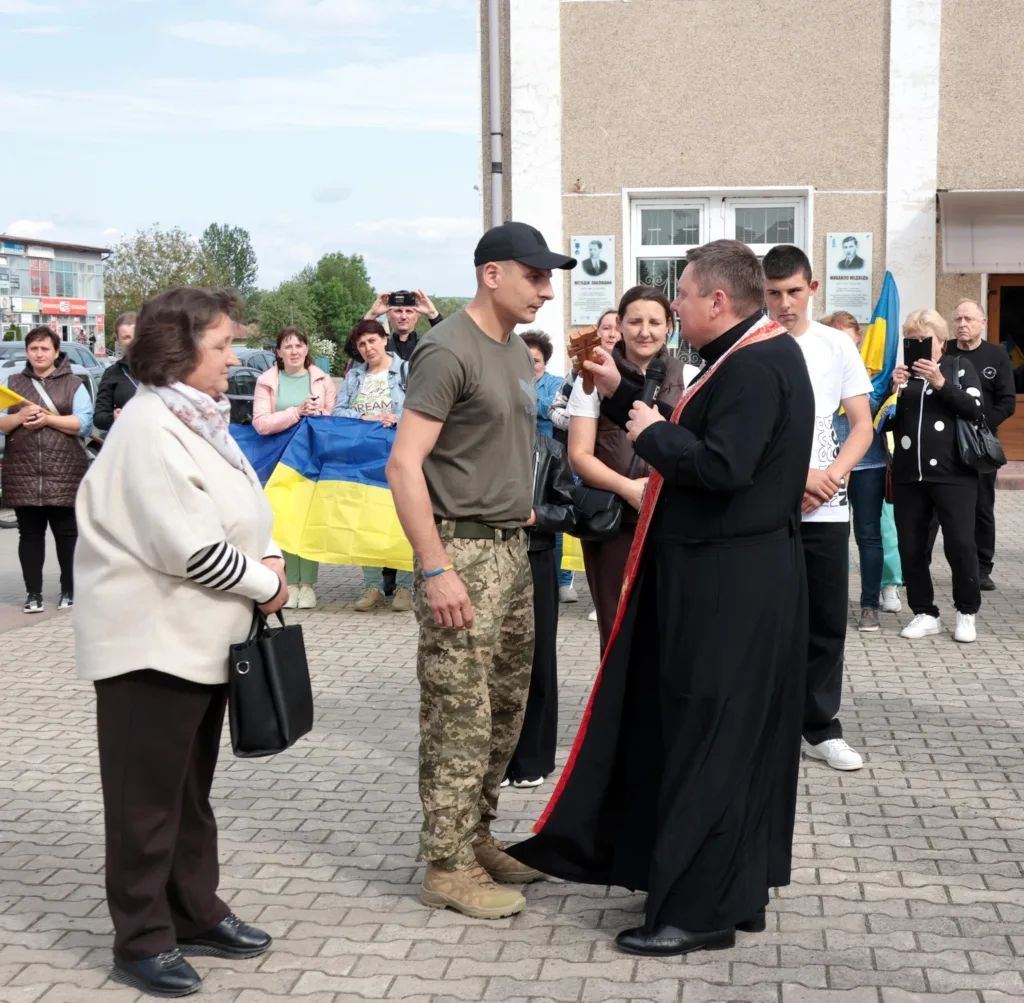 Священник благословляє військового перед людьми з прапором України