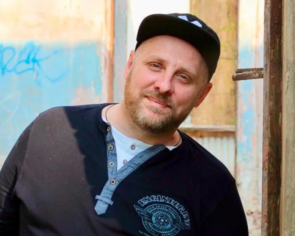 Чоловік у кепці посміхається біля старої стіни
