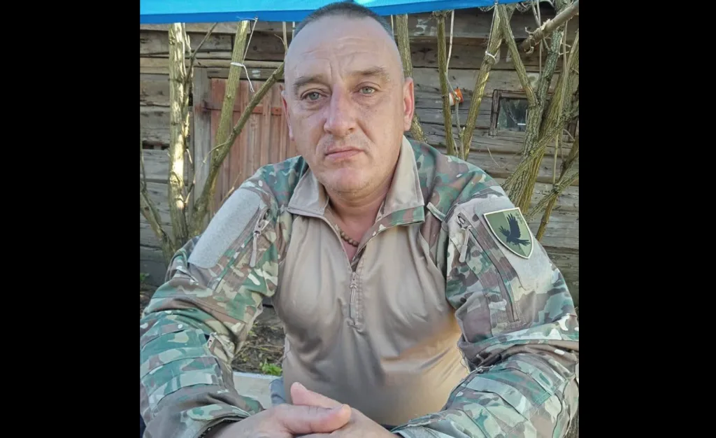 Чоловік у військовій формі сидить біля дерев’яного будинку