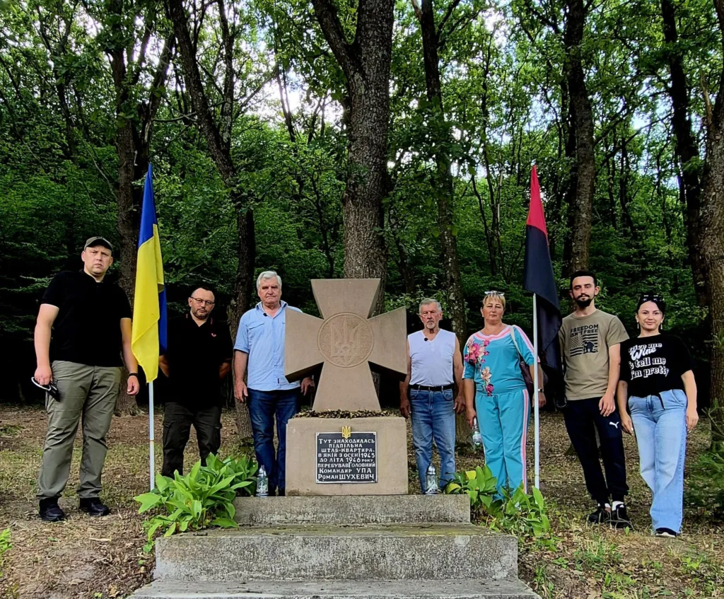Люди біля пам’ятника серед лісу з українськими прапорами