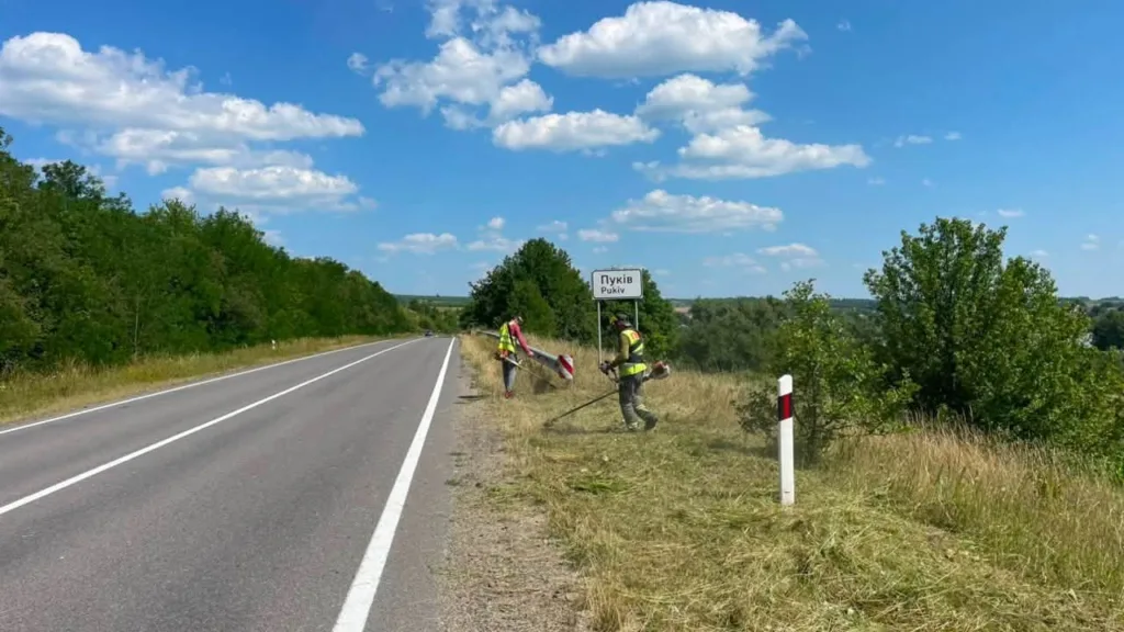 Робітники косять траву біля знаку села Пуків