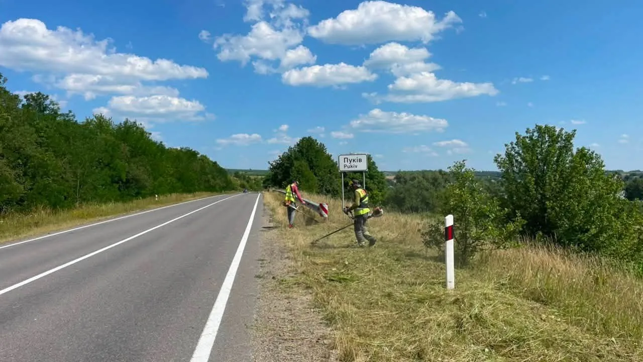 Робітники косять траву біля знаку села Пуків
