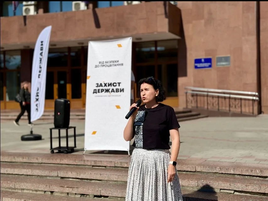 Жінка виступає з мікрофоном біля банера «Захист держави»