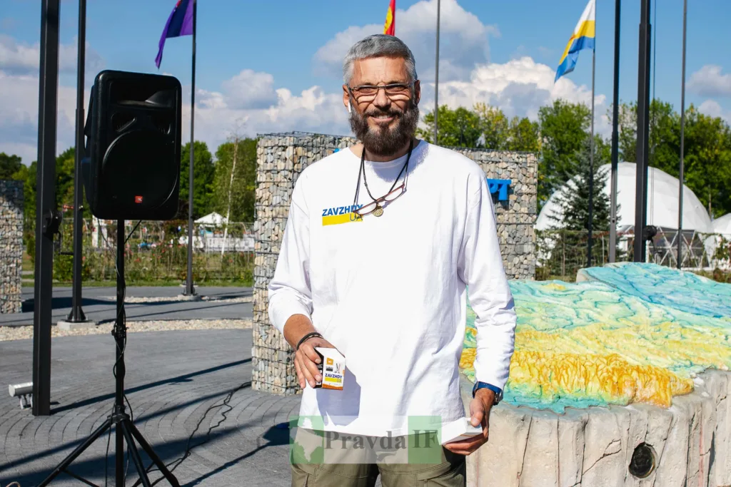 Усміхнений чоловік у футболці ZAVZHDY UA біля прапорів