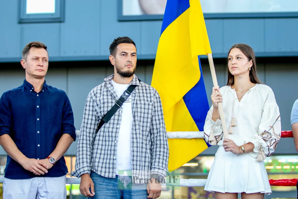 Троє людей стоять поруч із великим прапором України.