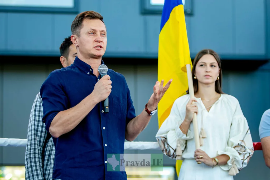 Чоловік виступає з мікрофоном поруч із українським прапором