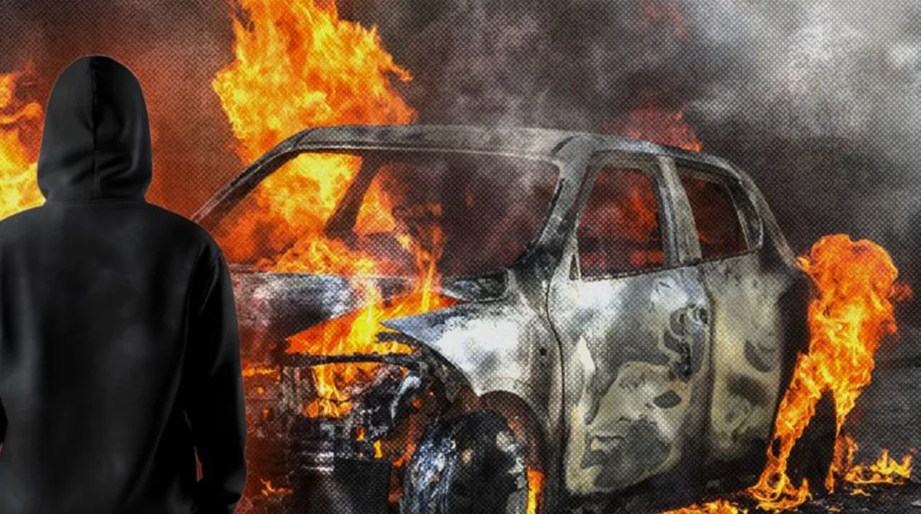 Людина в каптурі перед палаючим автомобілем