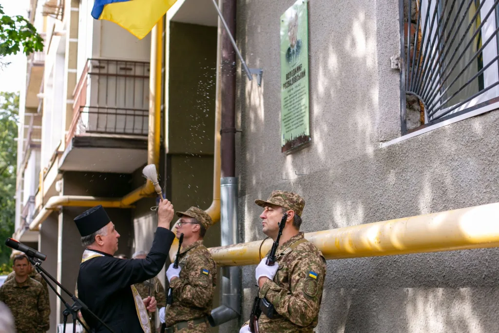 Священник освячує меморіальну дошку під українським прапором
