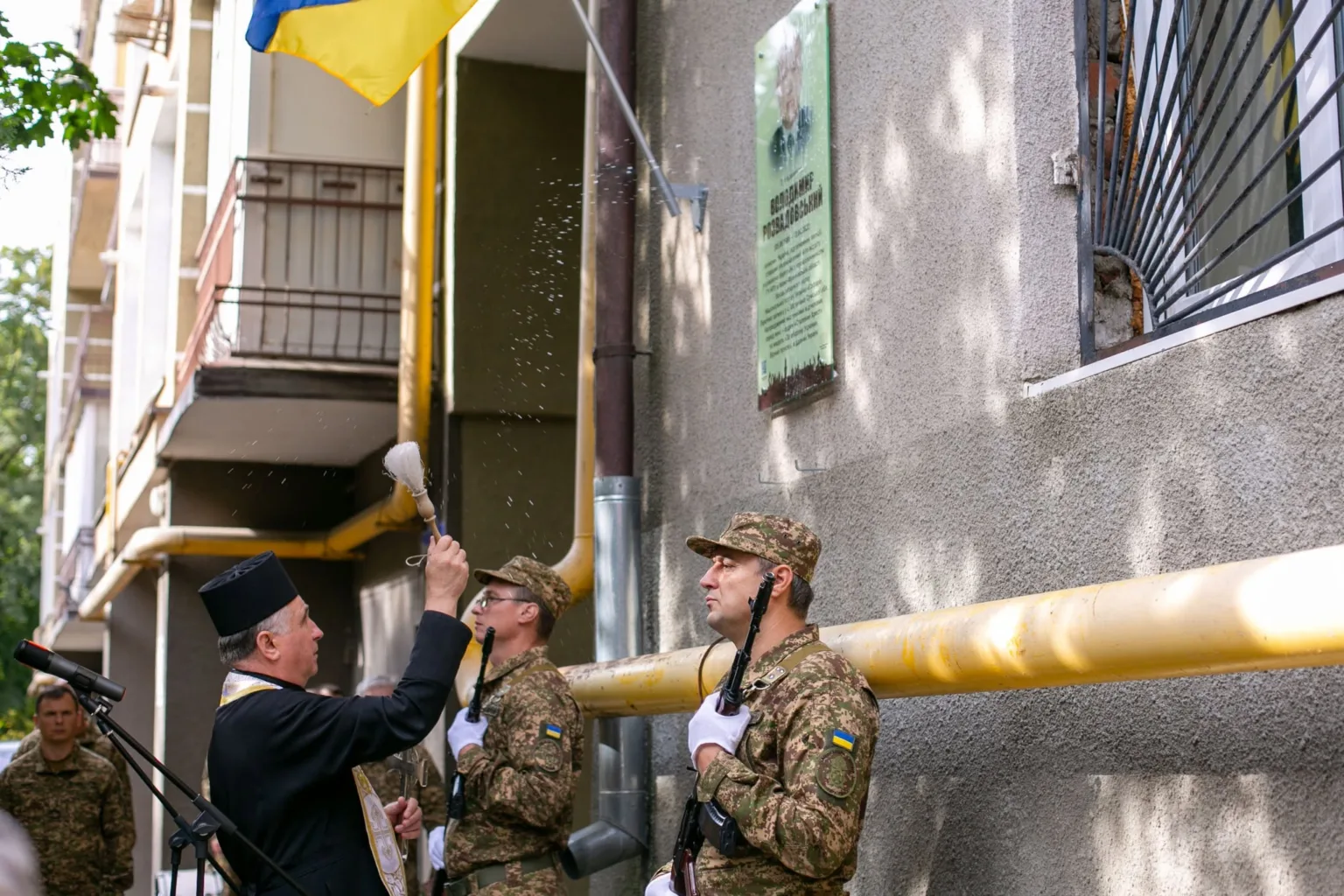Священник освячує меморіальну дошку під українським прапором