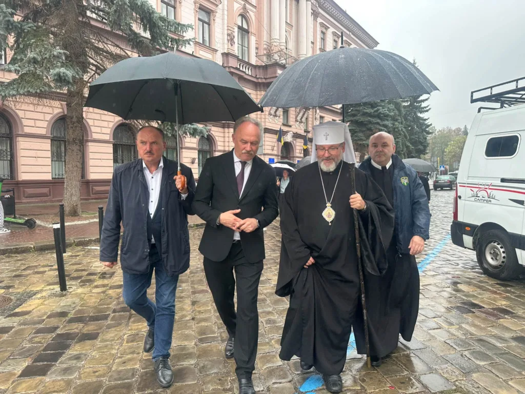 Священнослужитель і посадовці йдуть під парасолями під дощем