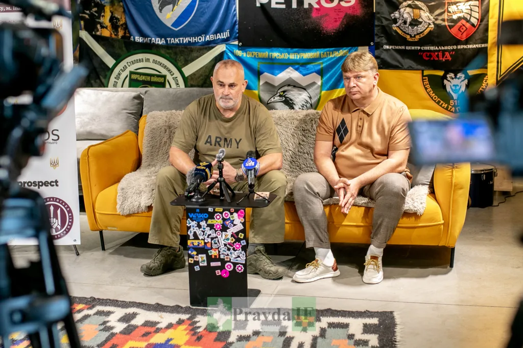 Двоє чоловіків дають інтерв’ю перед військовими прапорами