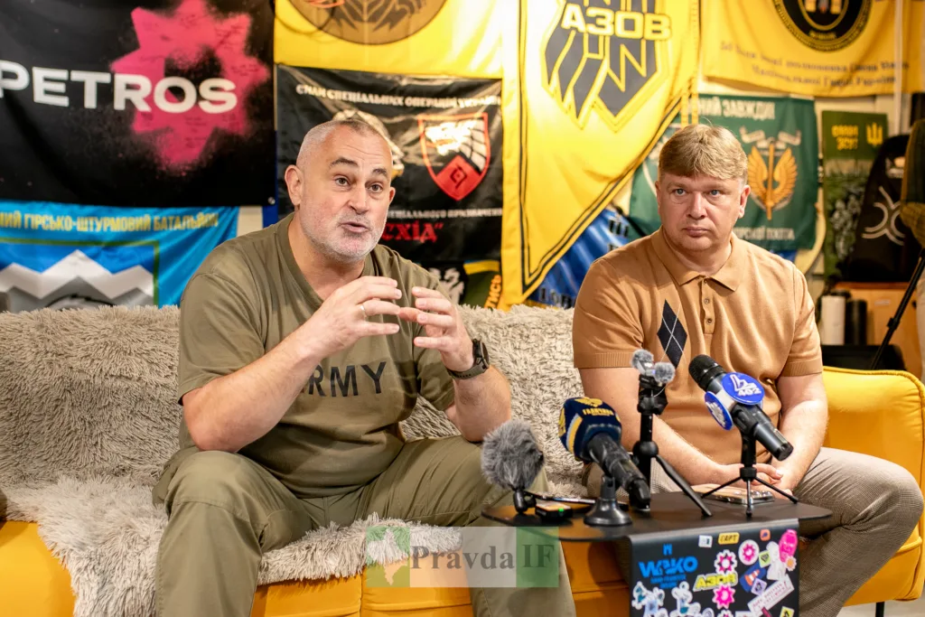 Двоє чоловіків дають інтерв’ю перед військовими прапорами