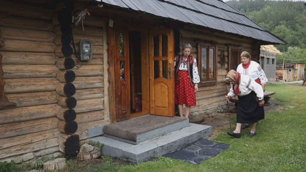Жінки в традиційному одязі біля дерев’яної хати